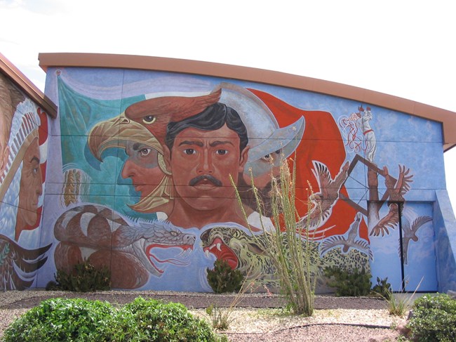 Three faces in front of Mexican flag: Native American in an eagle's mouth, Spaniard in metal helmet, and a Mexican man in the middle. Surrounding them are a serpent, jaguar, and the Papantla flyers.