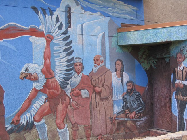 A Spanish priest stands with a Native American in front of a mission. A Spanish soldier sits under a tree while a Native woman stands next to him. A Spaniard holding a book stands to the right.