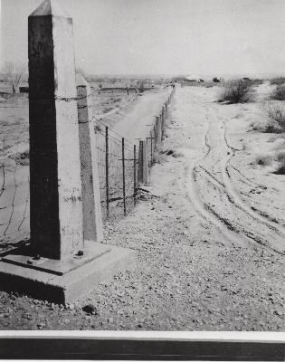 Black-and-white photo, obelisk monument in foreground followed by a line of concrete fence posts extending into background. All are connected by wire mesh fencing.