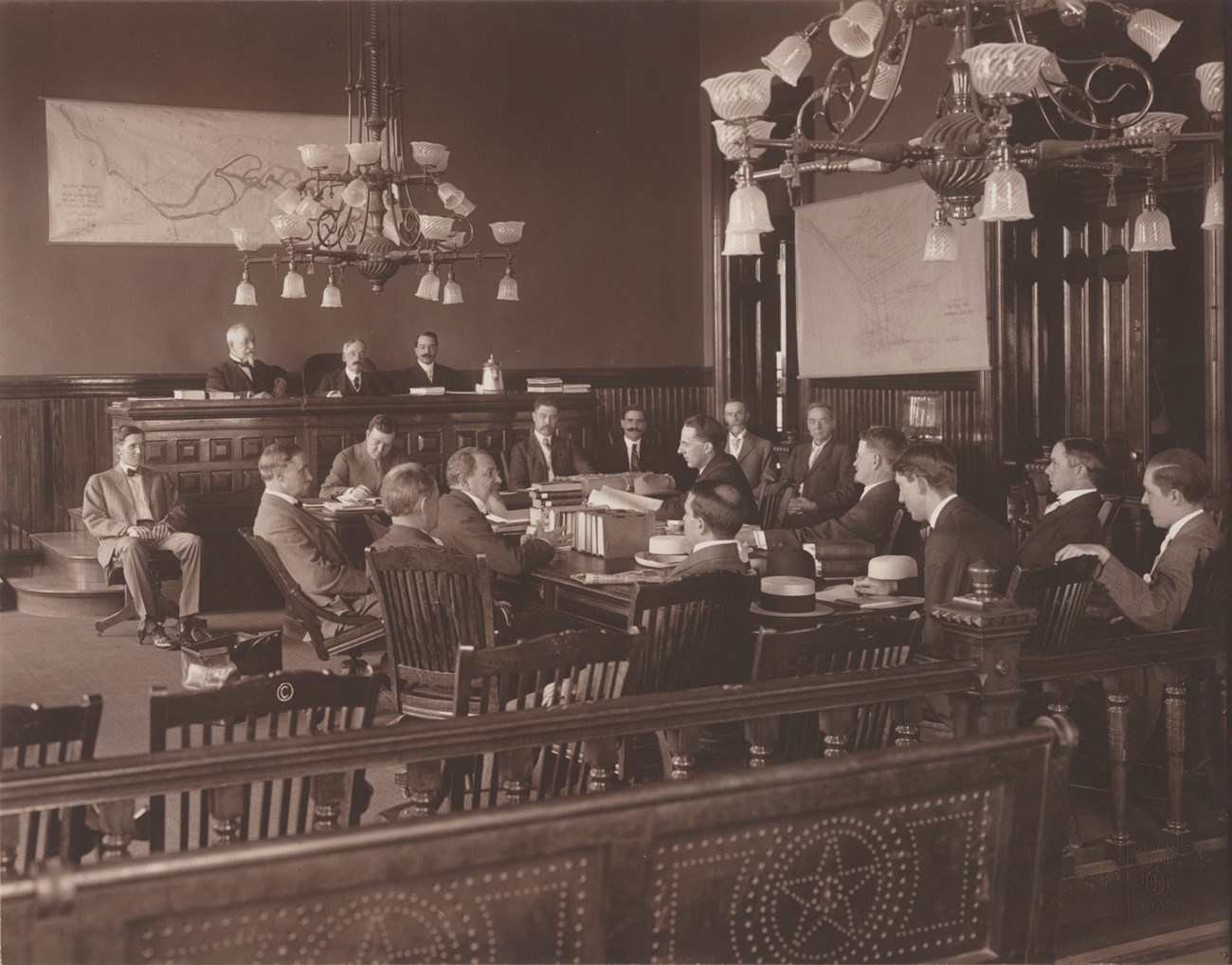 Black-and-white photo of 15 men around a table in an ornate room with paper maps posted on walls. Three commissioners sit behind elevated courtroom bench at far end of room.
