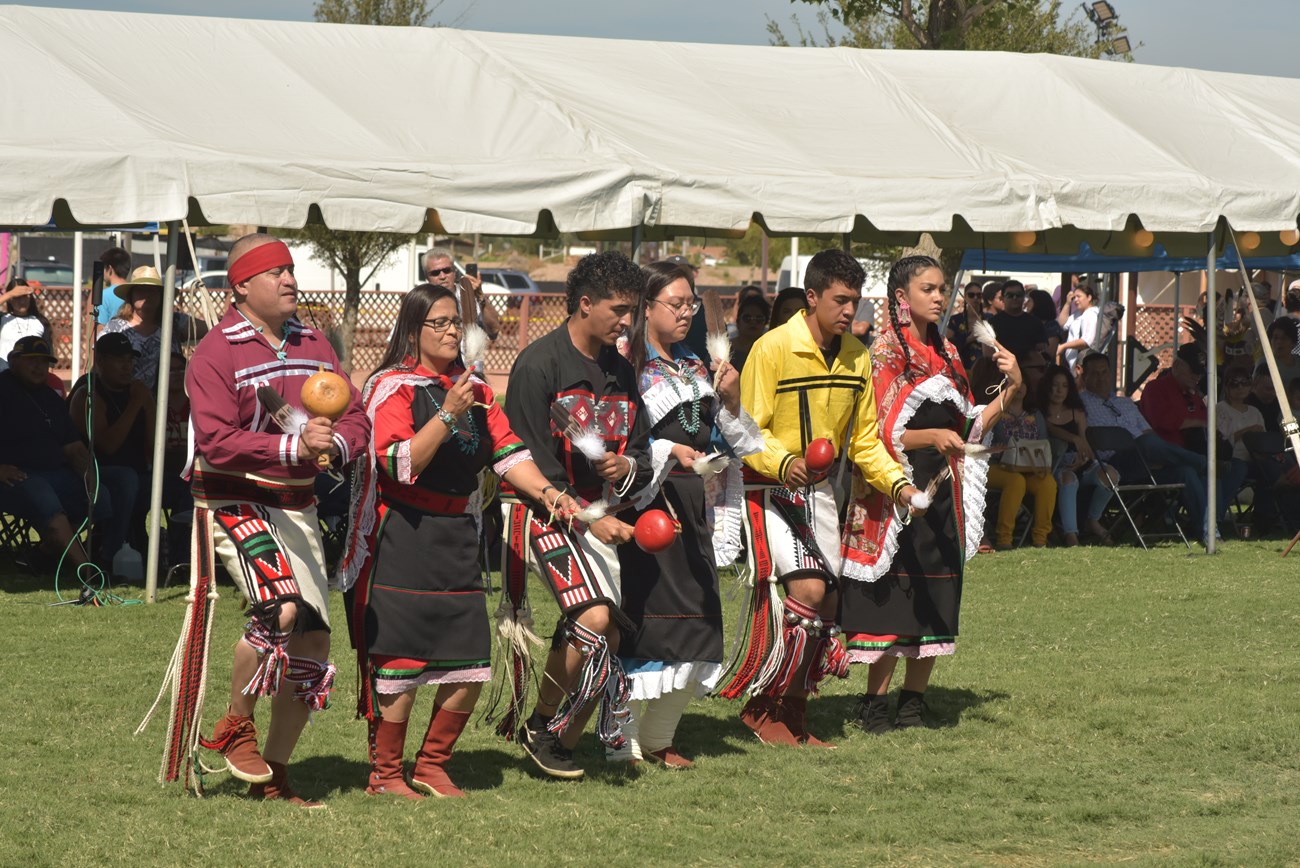 Three men and three women, dressed in traditional attire, dance side by side using gourd instruments and feathers.