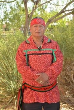 Man wearing a wide, red, patterned headband; a red, patterned shirt with green and black ribbons across his chest; and a red, black, and green patterned sash around his waist.