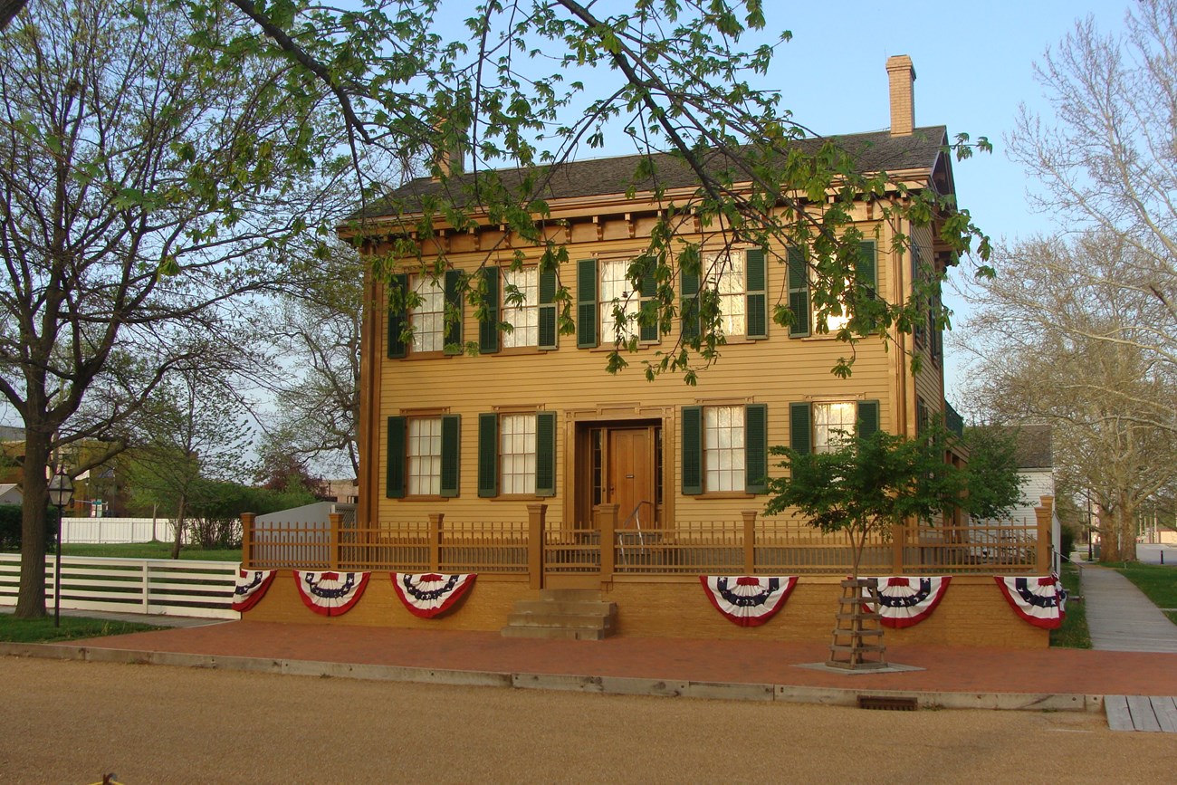 Tan, rectangular, two-story house with five green-shuttered windows on the top floor and two on either side of front door on the first floor. Fence around the small front yard is garlanded with semicircle flag banners.