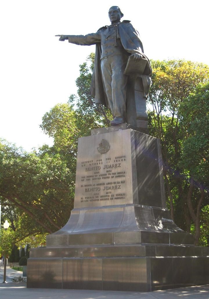 Bronze statue of a cloaked man holding a book in left hand and pointing straight ahead with right hand. Statue is mounted on a large, inscribed metal base.