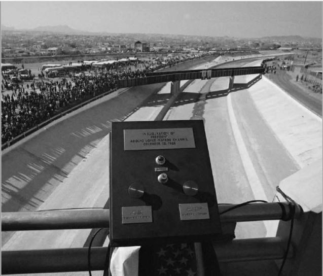 A black-and-white photo of a black box with labels and buttons mounted on the railing of a bridge overlooking a concrete river channel. A crowd lines the edge of the channel on the left side of the photo.