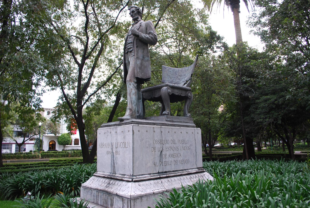 Bronze statue of a man standing on front of a chair is mounted on a large stone base inscribed with text. The statue is in a grassy area surrounded by trees.