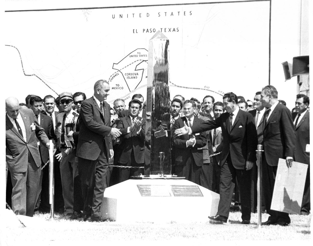 Two men surrounded by crowd of people walk toward each other with hands extended. A chrome obelisk monument is slightly behind them in the center of the scene.