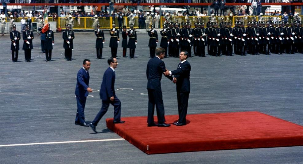 Two men shake hands on a red carpeted platform on airport tarmac surrounded by large crowd of people