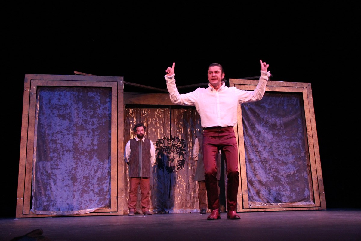Actor in brown pants and white shirt with hands raised stands near front of stage. Another actor stands in the background near simple set of curtain-covered doors.