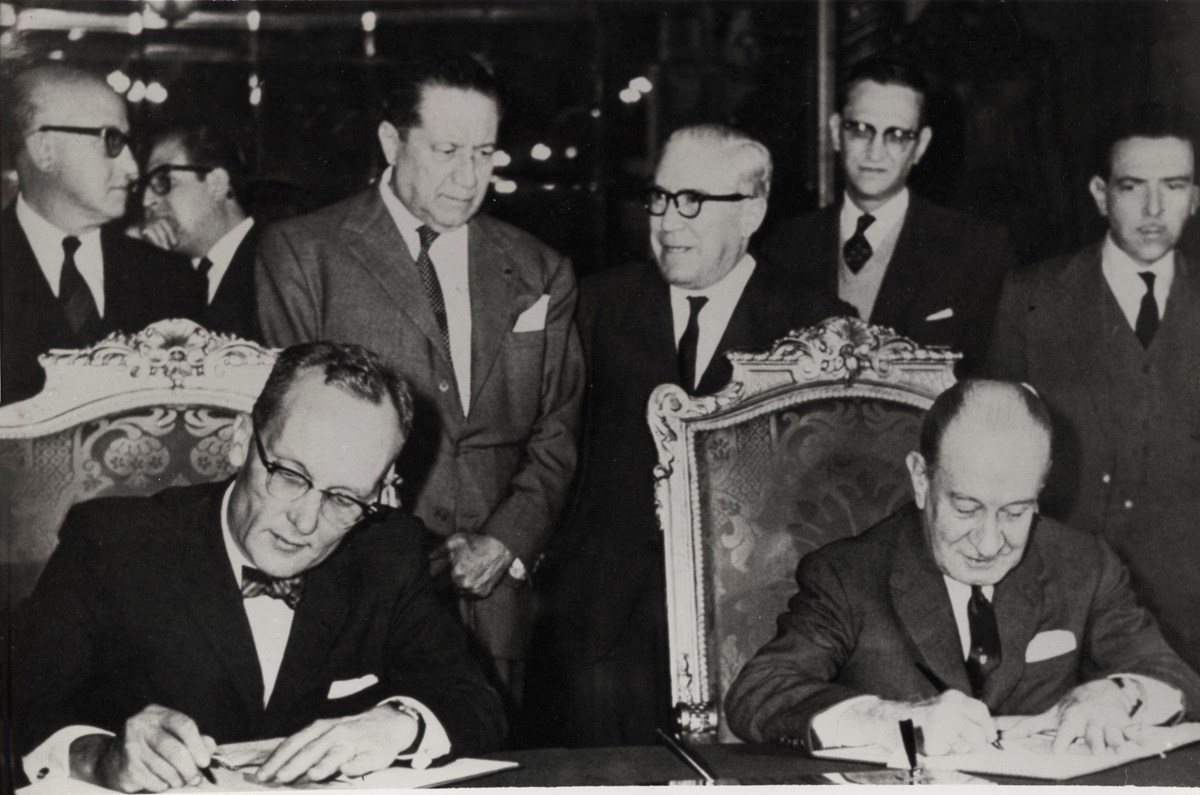 Black-and-white photo of two men in suits seated at table signing documents. Six men stand behind them.