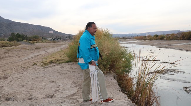 American Indian man wearing a blue shirt and white sash around his waist stands along the sandy banks of a low-flowing river. Browning vegetation lines the river banks. A rising plateau can be seen in the distance.