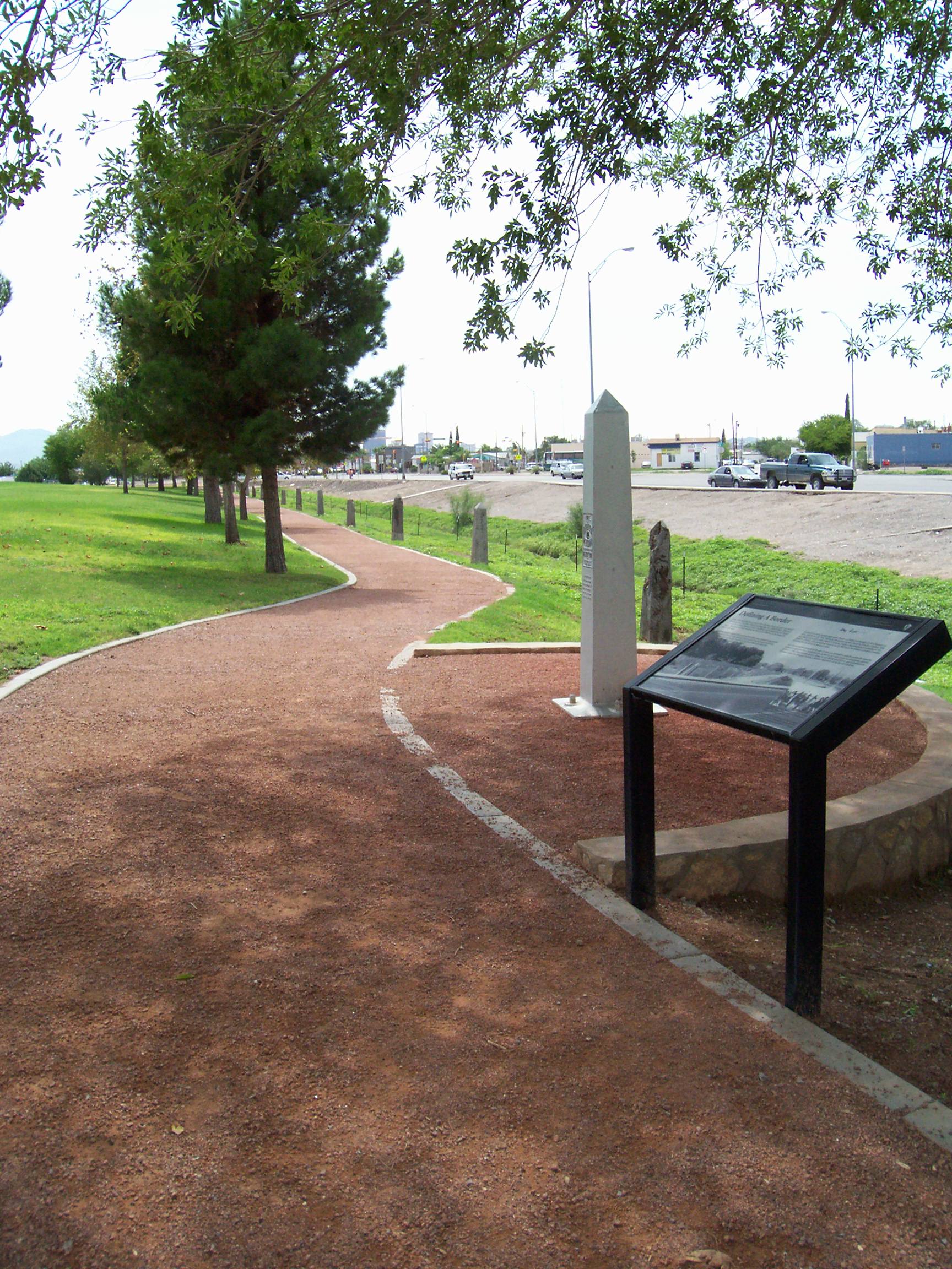 Boundary Monument Wayside Exhibit An interpretive panel stands 3 feet high next to a white obelisk, both along the edge of a wide, red gravel trail