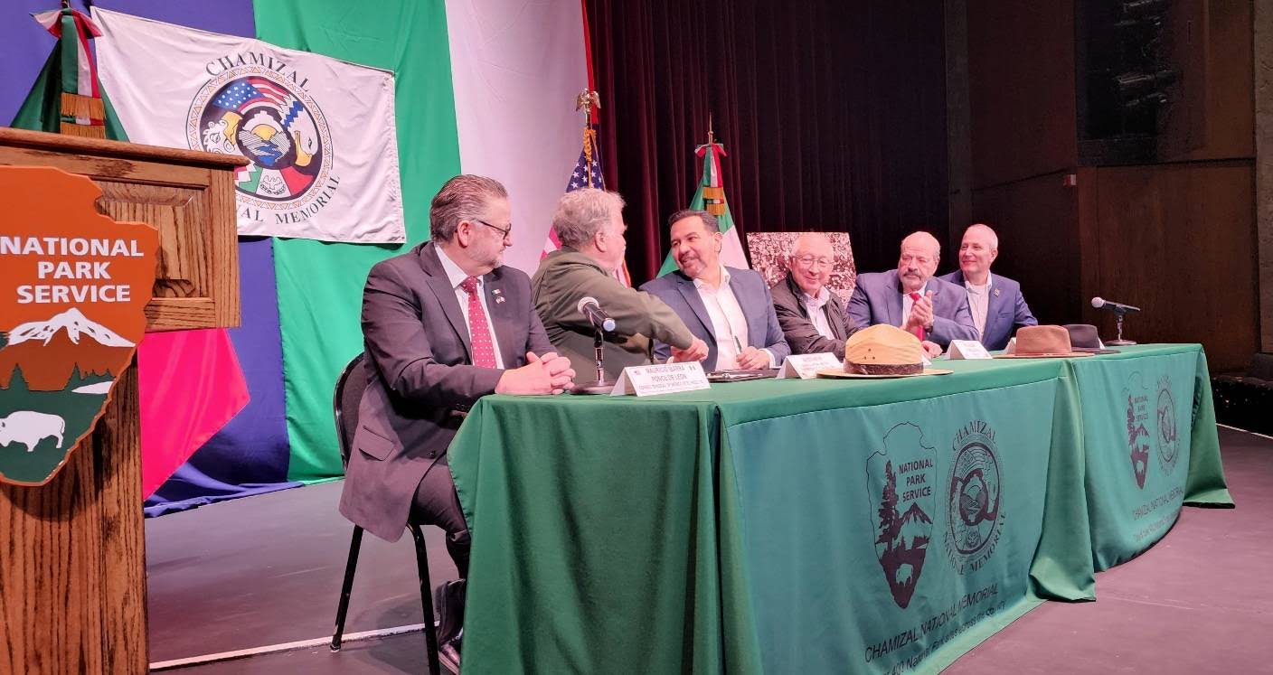 Six men sit at a long table on a stage. On your left side part of a podium adorned with the NPS arrowhead logo is in the image. A flag with Chamizal logo hangs on drapes behind the table, and US and Mexico flags are on the right. Two men shake hands.