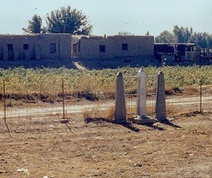 Shiny obelisk-shaped monument stands between two cement fence posts, adobe buildings in a field on opposite side of fence