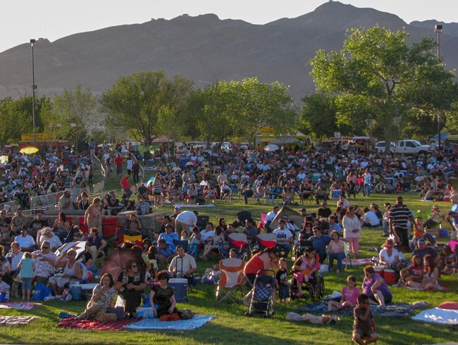 A crowd of people sit on a lawn in front of a distant row of trees, a mountain in the background.