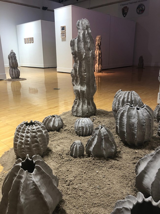 Ten cactus shaped sculptures sit in a bed of sand on the floor. Across the room are similar sculptures and framed art on a wall.