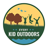 Round logo with a green field with two brown trees. Blue water feature in the foreground, three kids jumping in the field and three birds in flight.