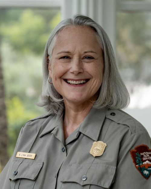 A smiling National Park Service ranger in a gray uniform with shoulder patch, badge, and name tag, standing indoors in front of softly blurred windows and greenery.
