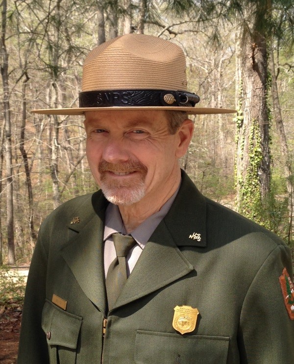 Bill Cox Retires A man smiling in a NPS uniform stands in a shaded tree lot.