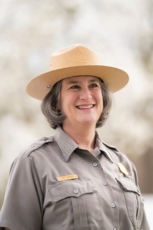 Standing woman light brown-grey hair smiles broadly while wearing her NPS uniform in front of blurred background.