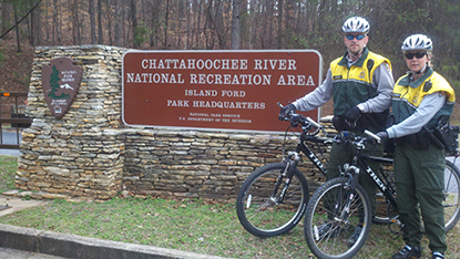 Two park rangers on bicycles stand next to a sign.