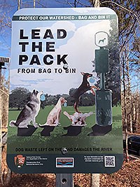 Sign showing 2 dogs waiting to drop their dog waste bags into the bin while a third dog is dropping its bag in the dog waste bin.
