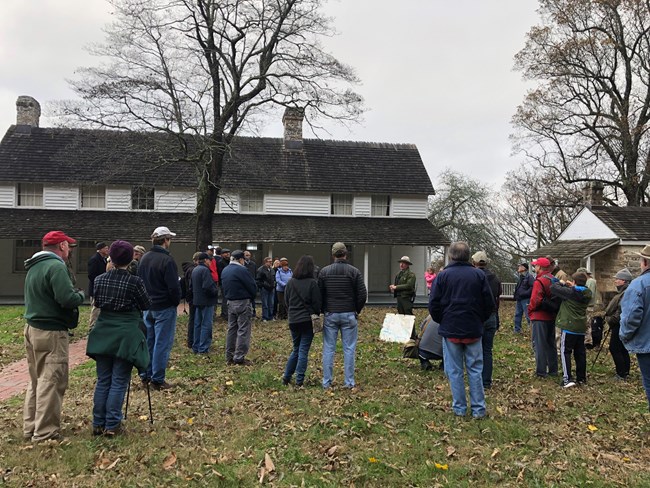 Park Historian Jim Ogden talks to a large group of people with the white planked Cravens House in the background.
