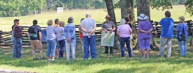 A ranger leading a battlefield overview tour. A ranger holding a map talks to a crowd of people in a field under a tree