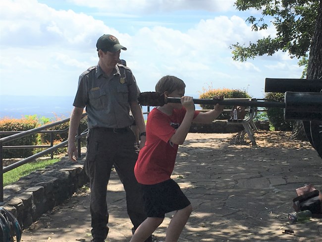 A male youth rams an imaginary cannonball down the barrel of a large cannon as a park ranger looks on.