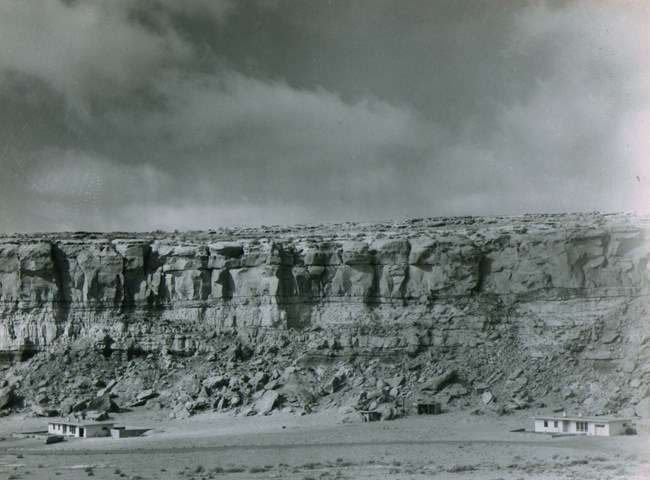 A black and white historic photo of 2 single story residences sitting at the bottom of a tall sandstone canyon wall.