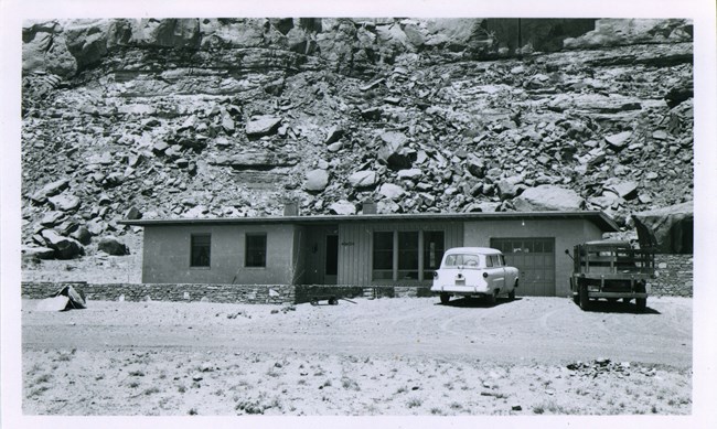 A black and white historic photo of a single story residence with canyon rock remnants in the background and cars shown parked in front.