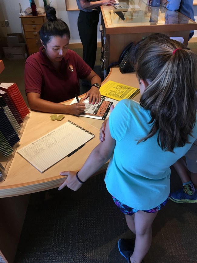 A woman in a red NPS shirt signing off on a completed Junior Ranger booklet at the front desk in the visitor center.
