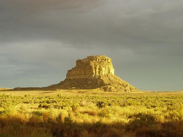 A tall sandstone butte sunlit in front of a gray sky.