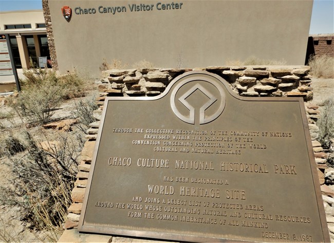 A brown sign designating Chaco Culture National Historical Park as a World Heritage Site sits in front of a tan building identified with text as the Chaco Canyon Visitor Center.
