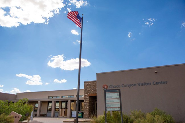 An adobe building with the words "Chaco Canyon Visitor Center" written next to the National Park Service arrowhead, with an American flag flying on the flagpole in the foreground.