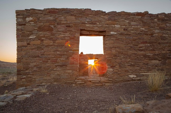 The sun rises through two doorways of remains of a very detailed masonry structure.
