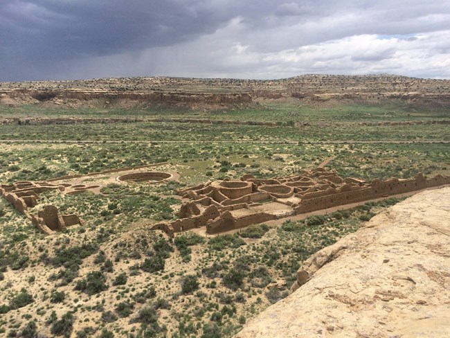 Remains of a large masonry structure with open rectangular and circular rooms rises above the desert. Looking down on the large building from above, storm clouds can be seen in the distance.