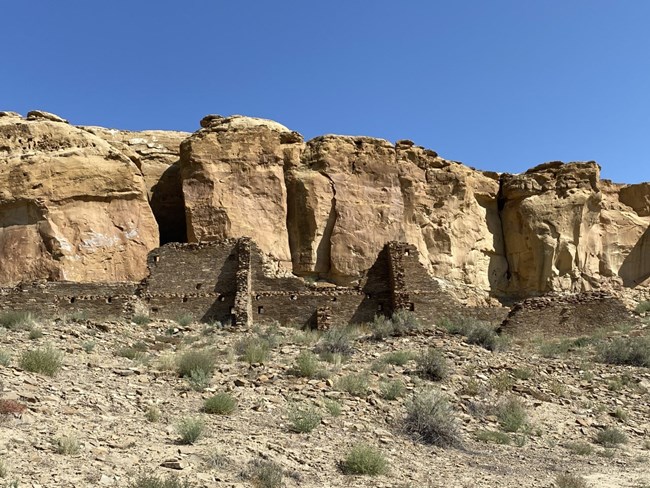 Remains of brown masonry walls reaching one to two stories above the desert floor, with a tall canyon wall just behind the unexcavated site.