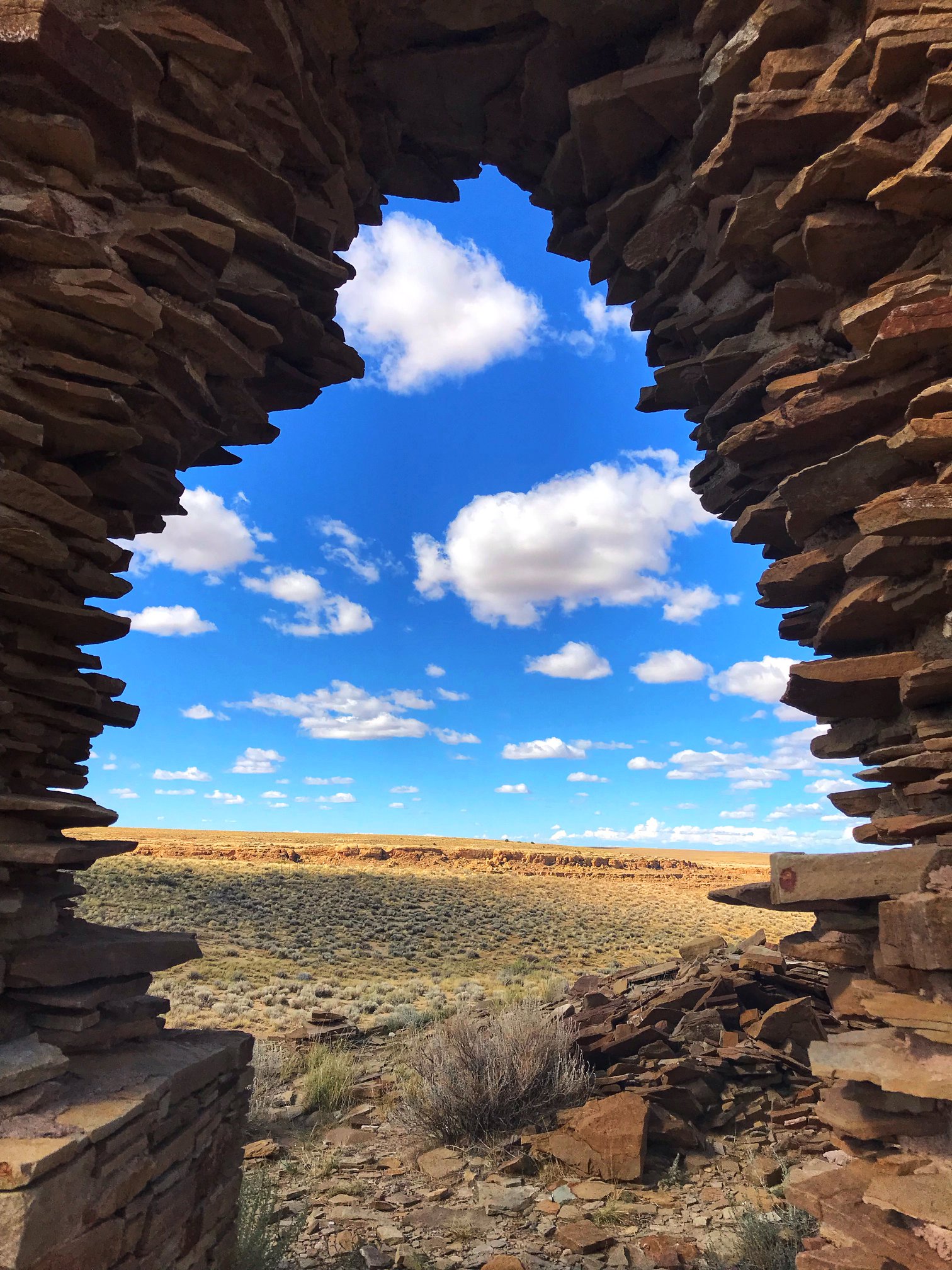 A tall masonry wall with a jagged doorway acts as a window to show the remains of masonry walls in the background, mixed among and blending into the desert landscape.