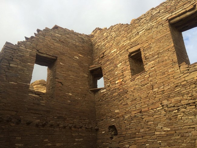 Two walls with extremely detailed masonry work rise above the viewer, with 4 doorways or windows visible - including a doorway in the corner of these two walls. Several logs from a previous ceiling are emerging from one wall to the bottom left.