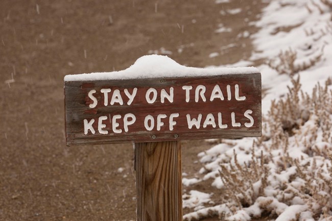 A wooden sign that says "Stay on Trail. Keep off Walls." with snow on the top.