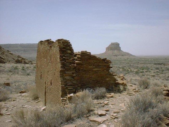 Remains of a large masonry wall emerging from the desert floor, with a tall butte in the background.