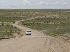 A vehicle drives on a wide dirt road through the desert.