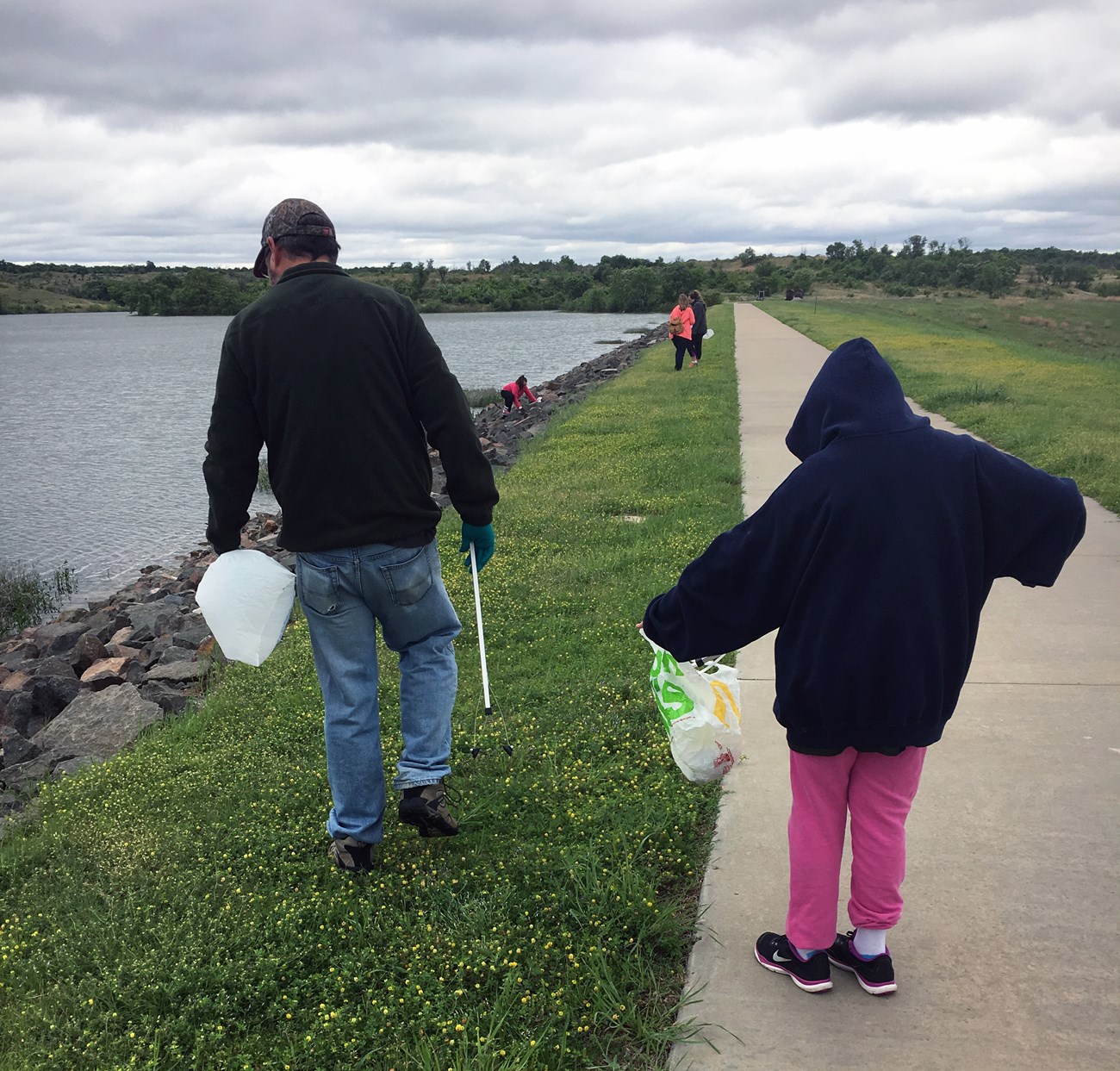 A man holding trash tongs walks along a sidewalk near a lake. To his right is a girl putting trash into a bag. Several more people are in the background picking up litter.