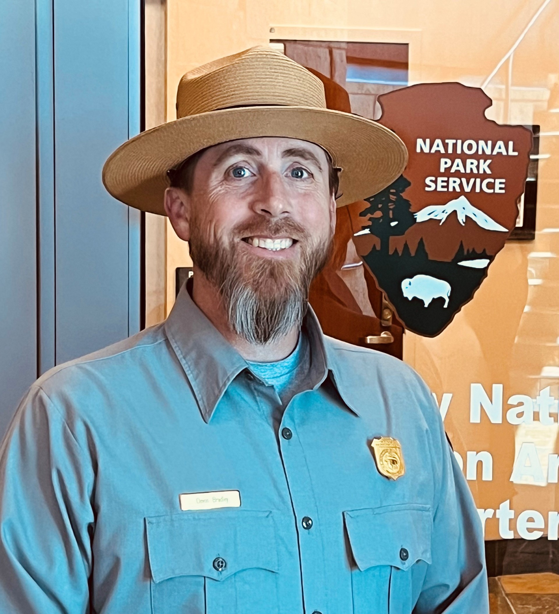 Bradley Bio Pic - NPS Photo a smiling man in a ranger hat and a grey uniform shirt stands near a park service arrowhead logo