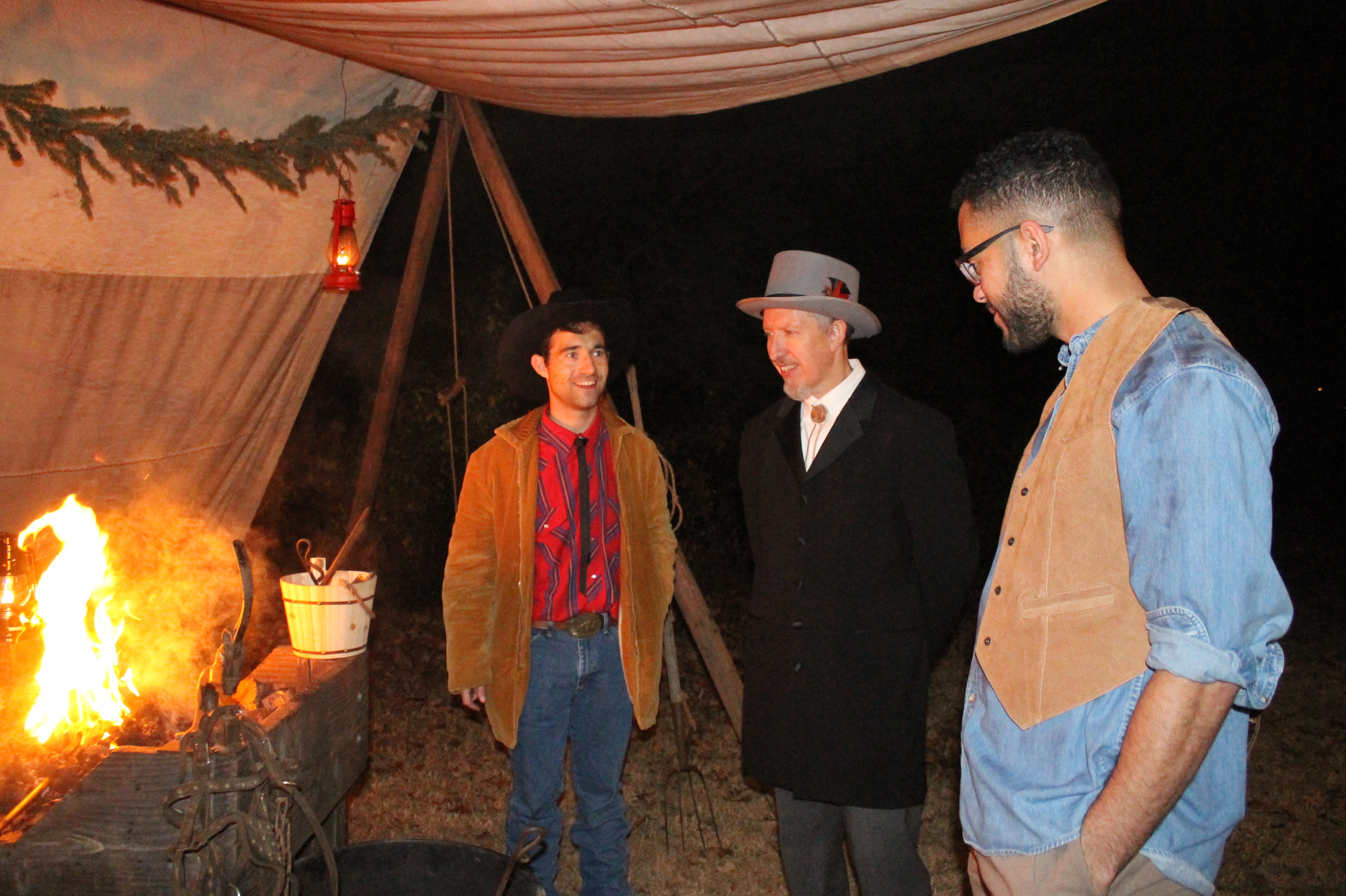 Three men in historical clothing stand near a fire.