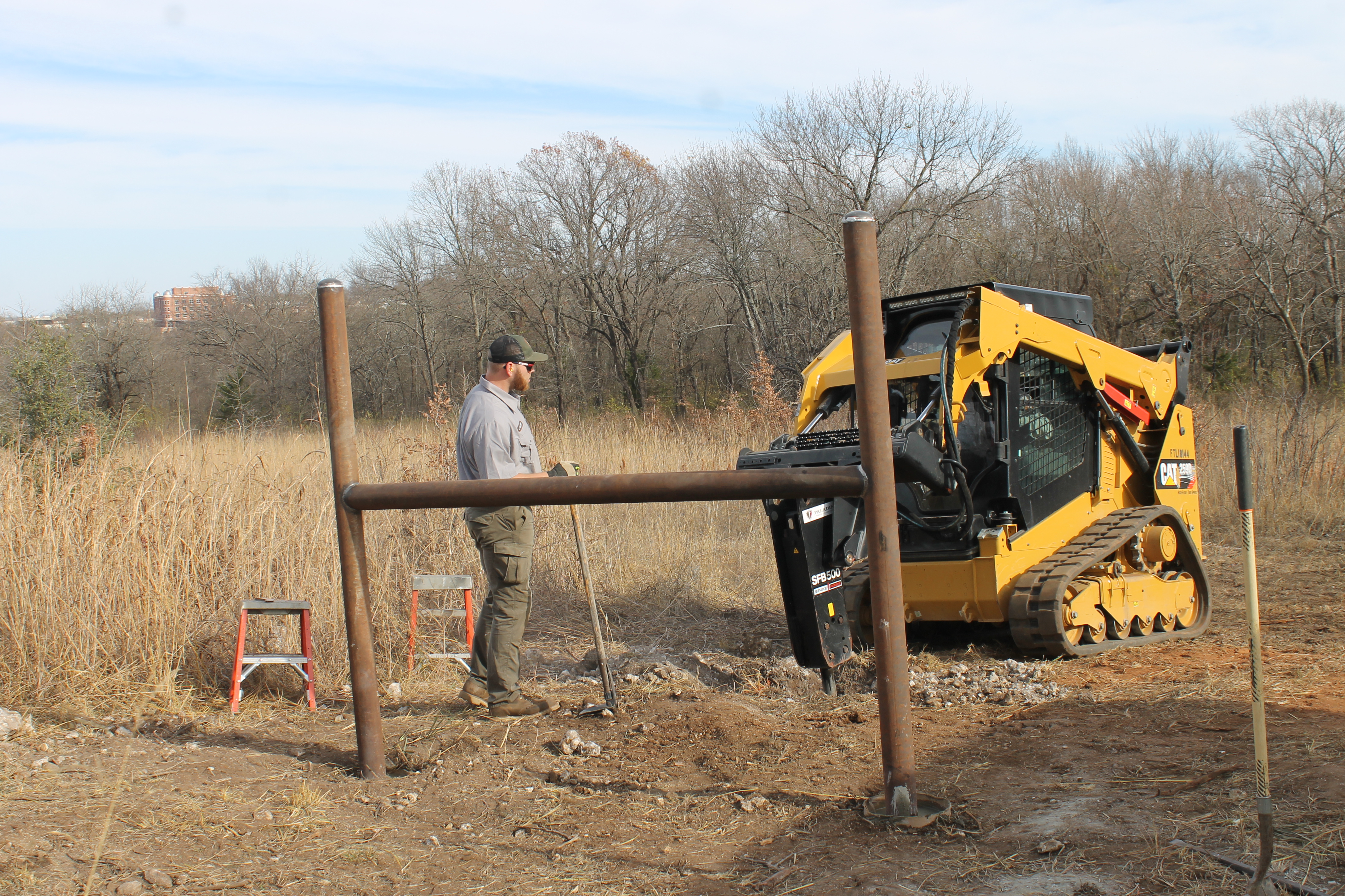 A worker with a shovel and a small tractor dig a hole next to a fence post.