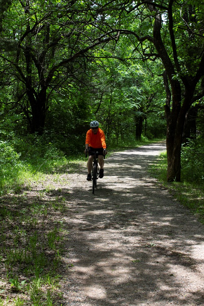 A man wearing a bright orange and a white helmet rides a bike on a gravel trail through a forest