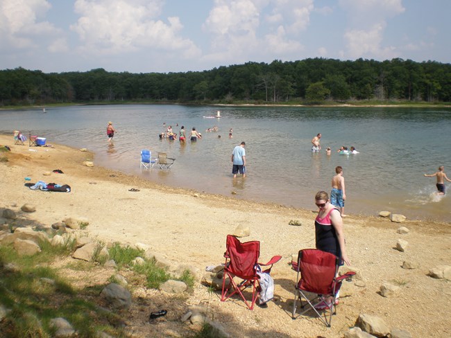 Several people swim in the water in a lake. Chairs and other objects sit on the lakeshore.