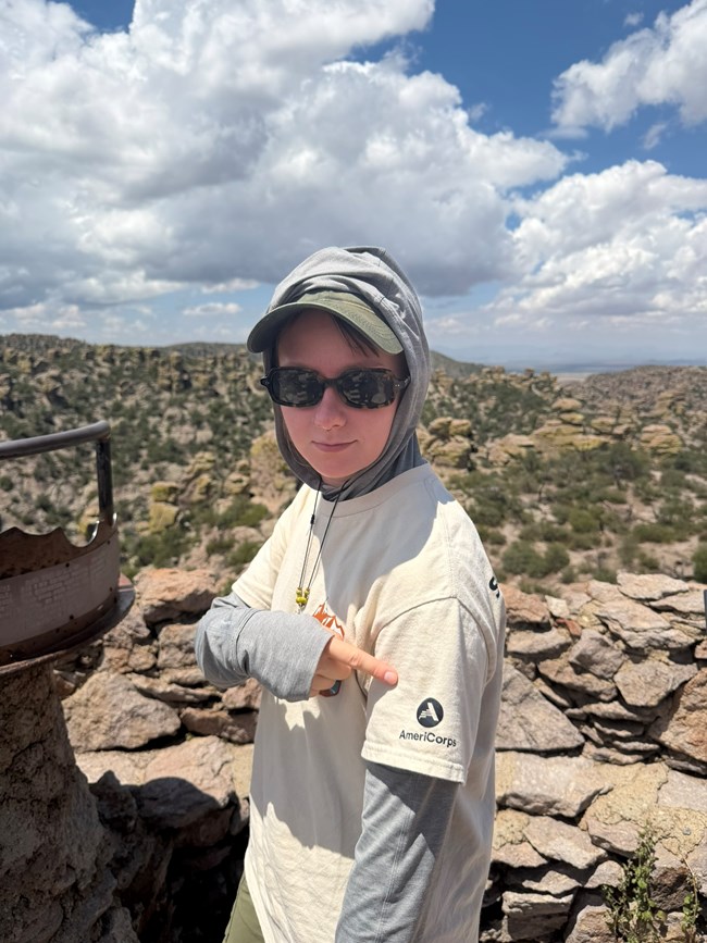 intern wearing an Americorps t-shirt stands at a lookout on Massai Point with rock formations all around